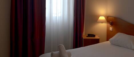 A hotel room with a bed, a nightstand, a lamp, and a window with curtains.