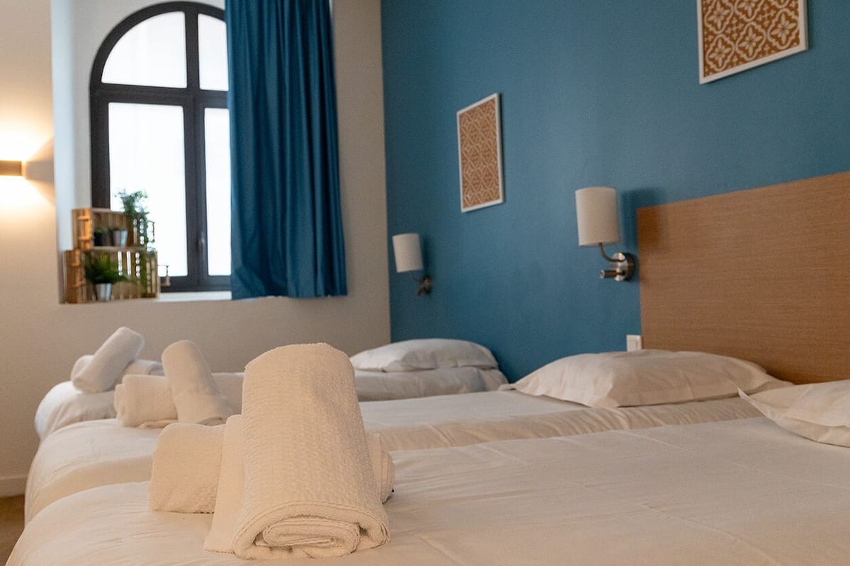 Een hotelkamer met twee bedden, een groot raam met blauwe gordijnen en decoratieve wandkunst.