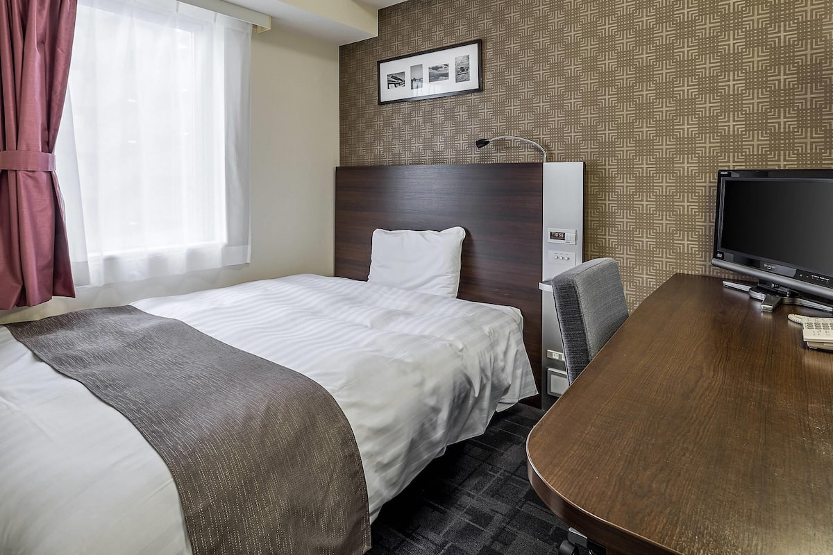 Quarto de hotel com duas camas, uma escrivaninha, uma televisão e um painel de controlo fixado na parede.