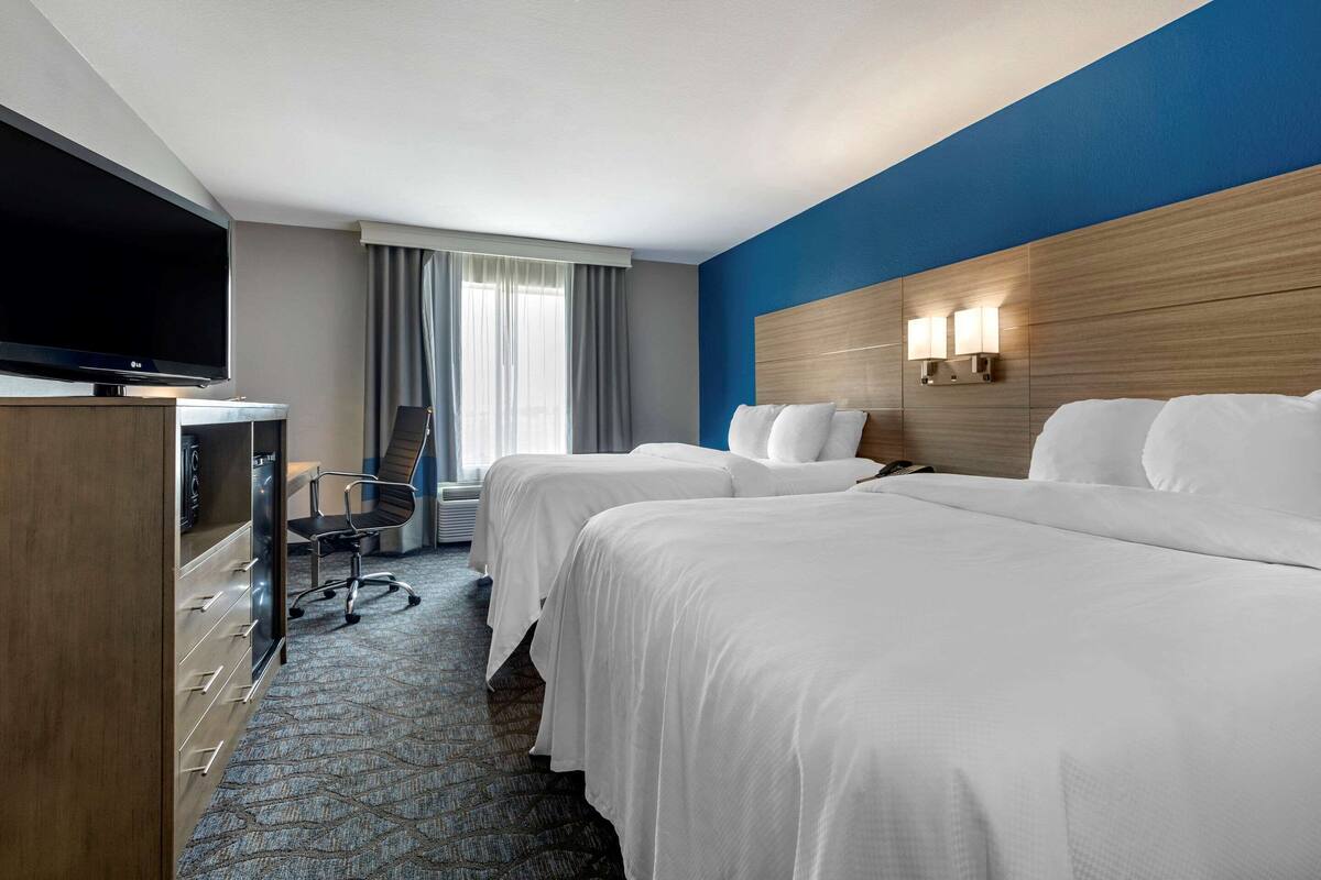 A hotel room with two beds, a TV, a desk with a chair, and a large window with curtains.