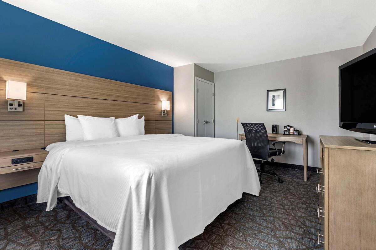 A hotel room with a large bed, a desk, a chair, a television, and a wall-mounted lamp.