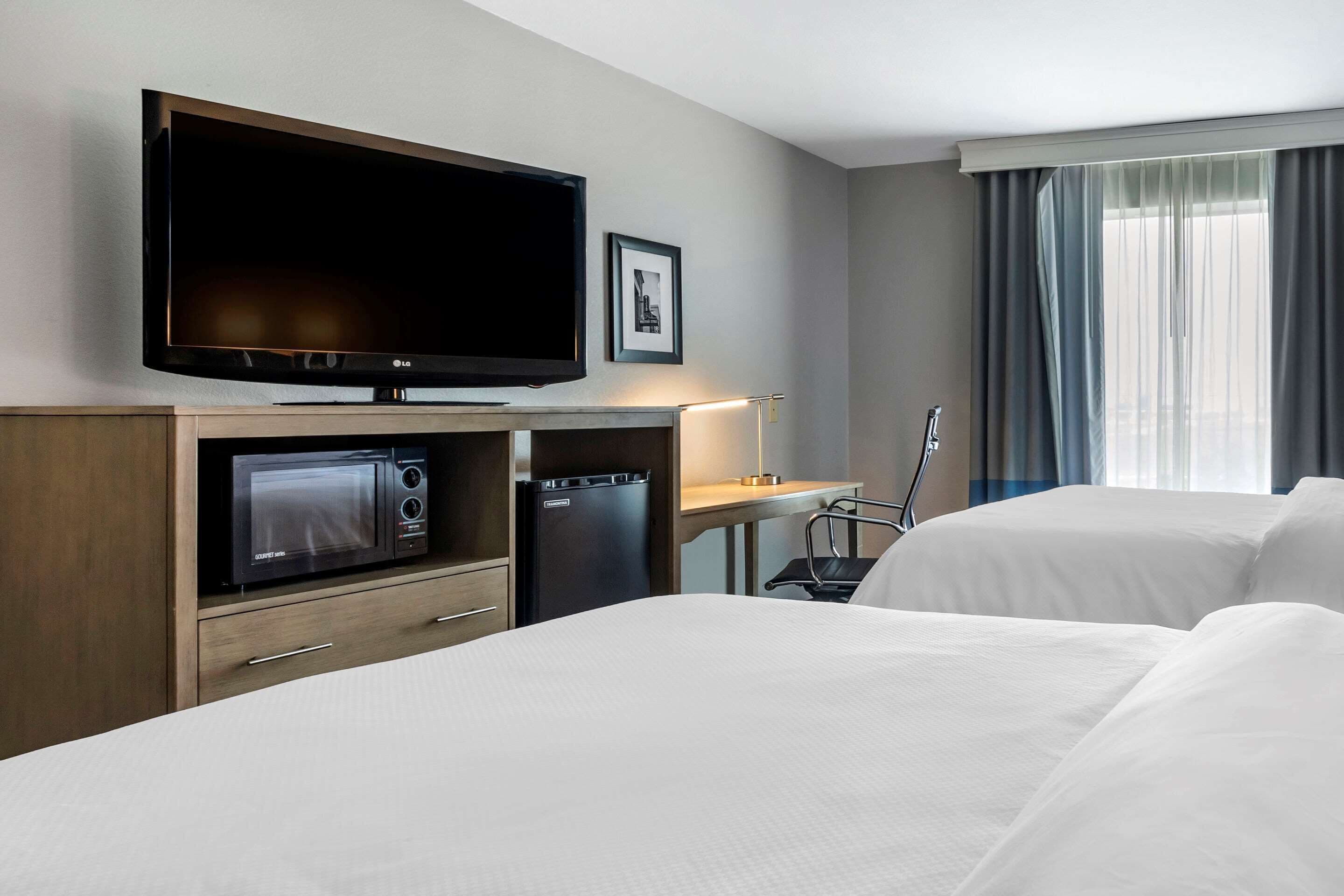 Habitación de hotel con televisor de pantalla plana grande, microondas, refrigerador minibar, escritorio con lámpara y cama con ropa blanca.