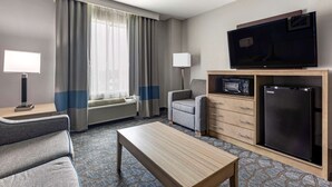 Une chambre d’hôtel équipée d’un canapé, d’une table basse, d’une télévision, d’un four à micro-ondes, d’un réfrigérateur, d’une lampe et d’une fenêtre avec des rideaux.