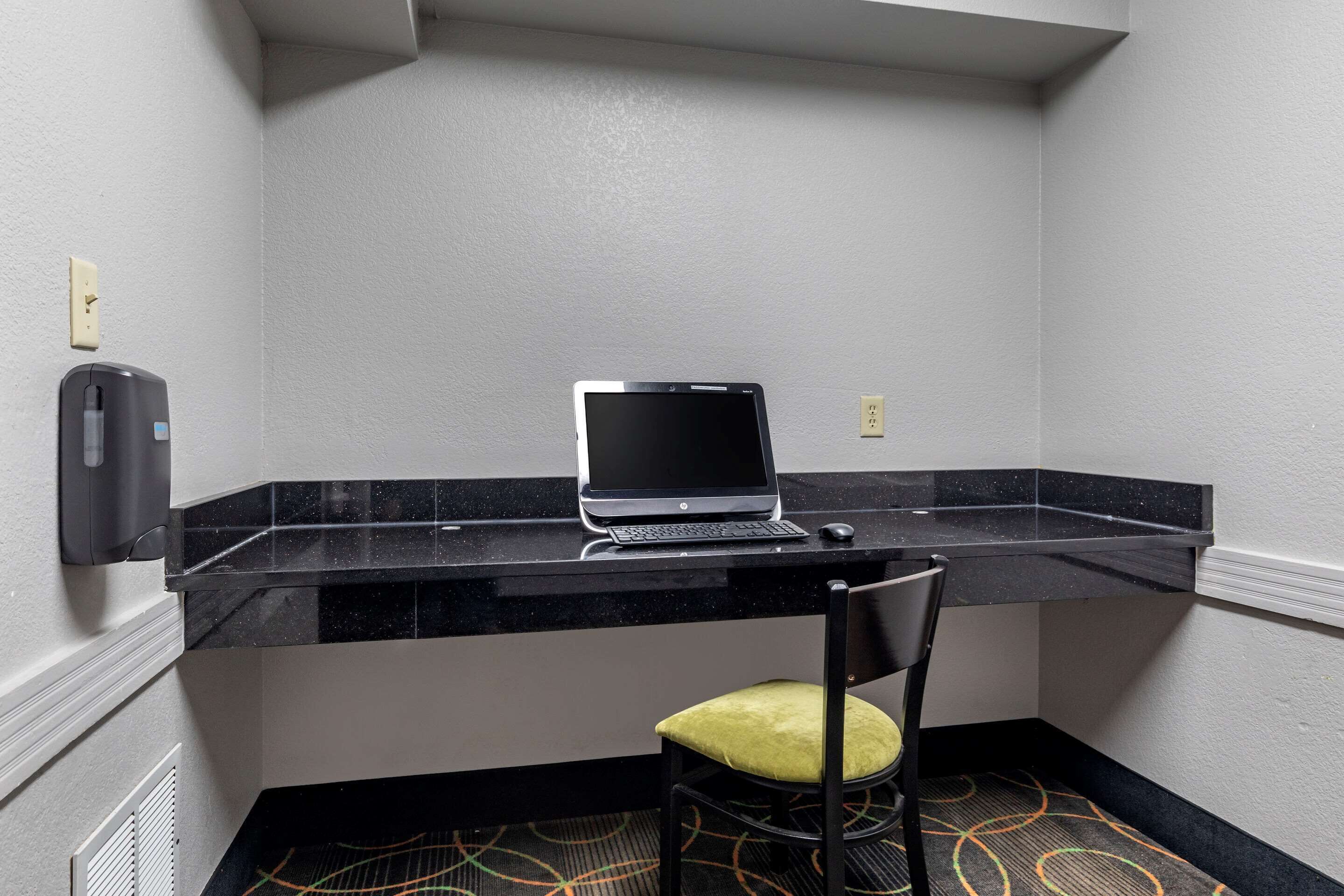 Un bureau d’angle avec un ordinateur portable posé sur un bureau en marbre noir, une chaise avec un coussin jaune et un distributeur de gel hydroalcoolique fixé au mur.