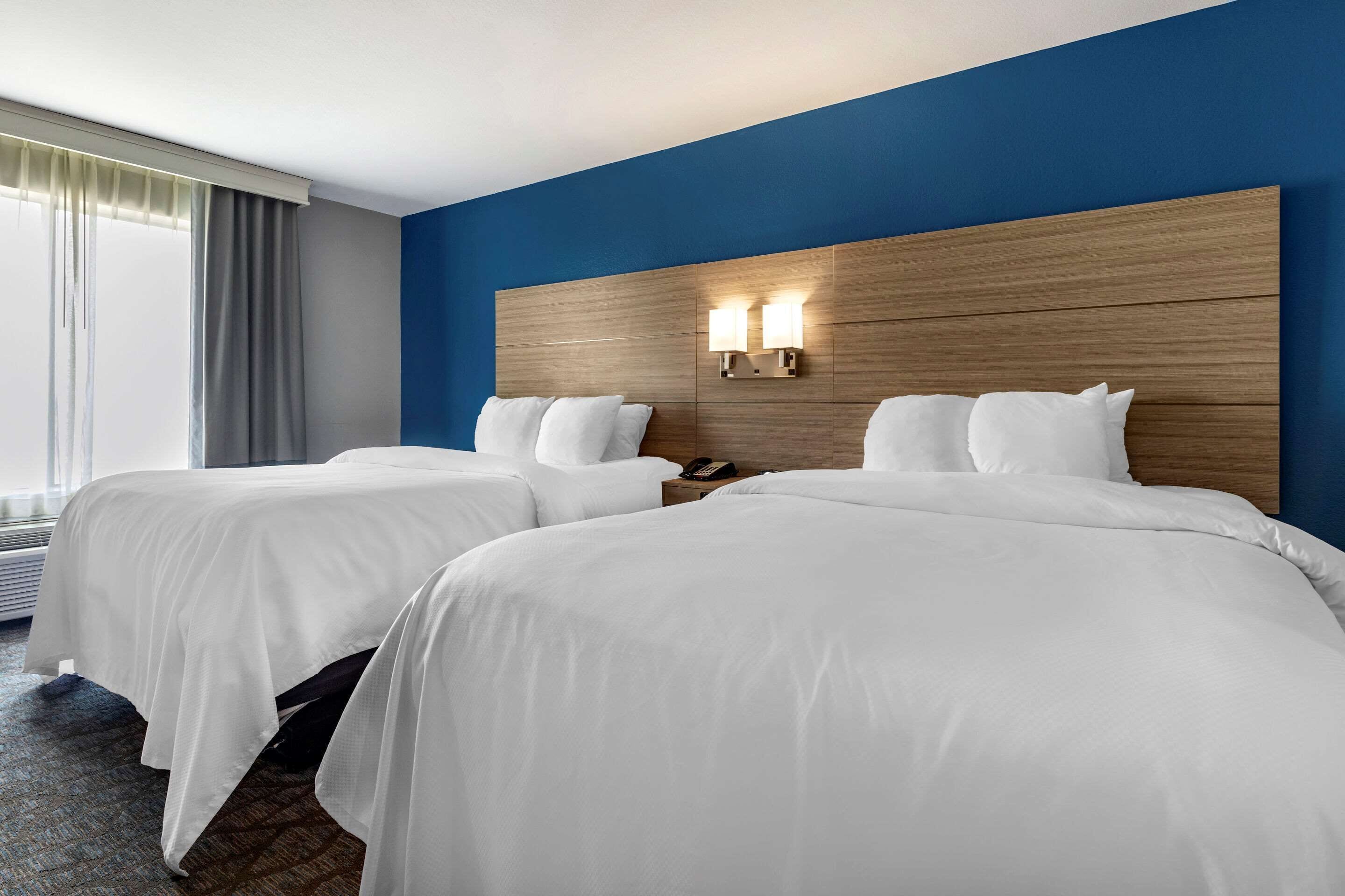 Quarto de hotel com duas camas, uma cabeceira de madeira e duas luminárias fixadas na parede.