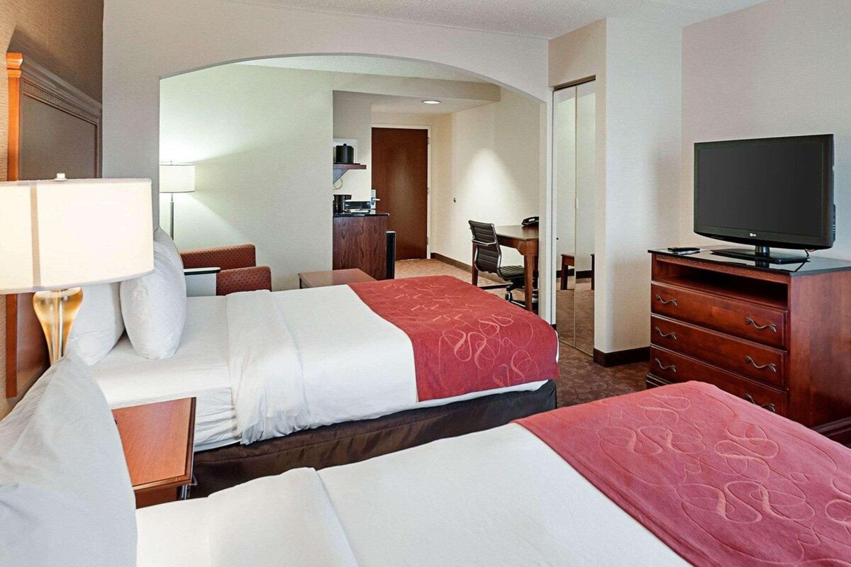 A hotel room with two beds, a desk, a television, and a small kitchenette.
