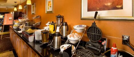 Un bufé de desayuno de hotel con una máquina para hacer gofres, una cafetera y diversas opciones para el desayuno.