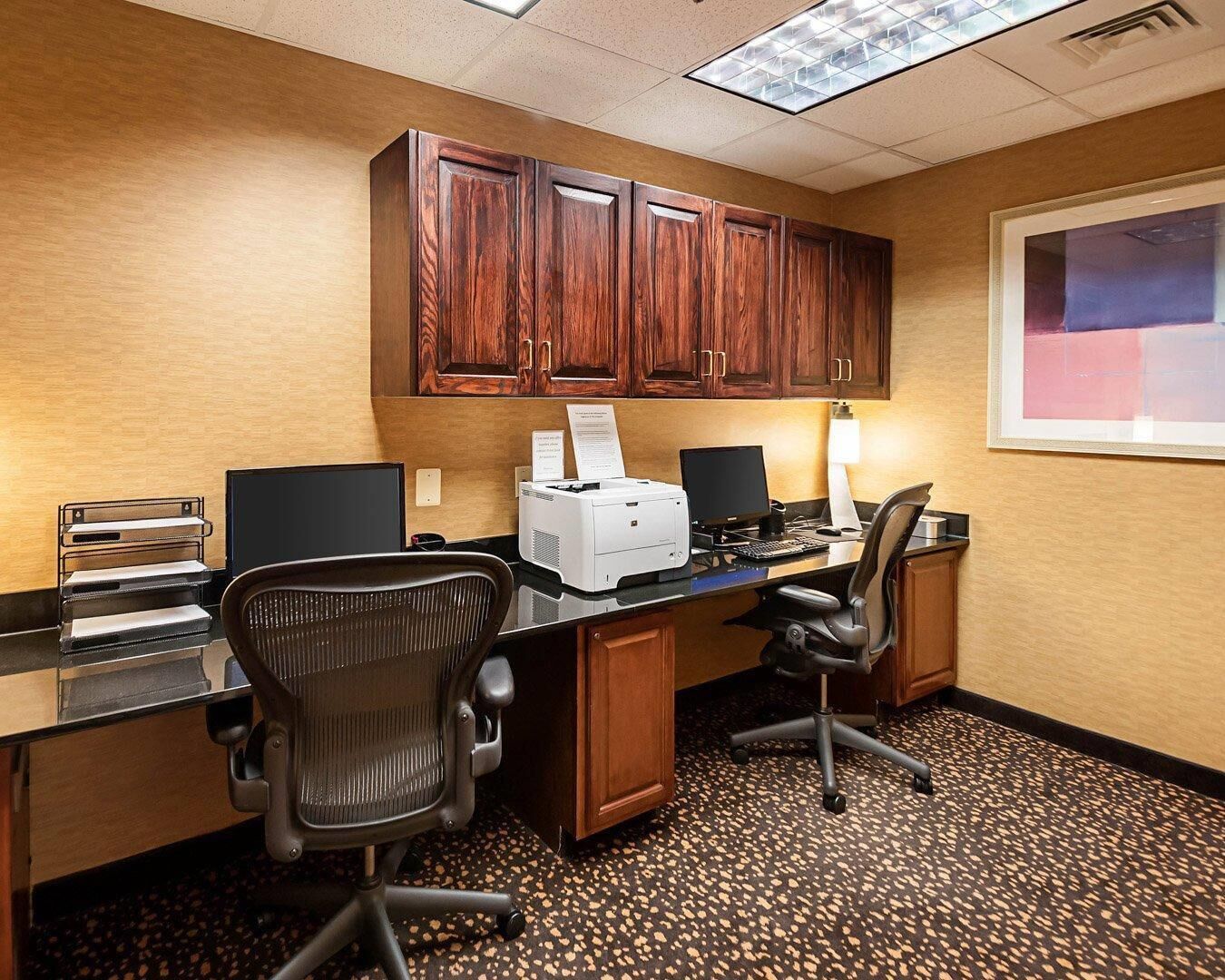 A hotel office with a wooden desk, two chairs, a printer, and a computer monitor.
