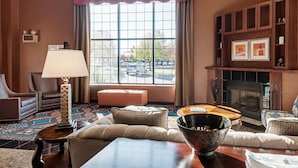 A living room with a fireplace, a large window, a sofa, a coffee table, and a lamp. - Comfort Suites Dulles Airport (Chantilly)