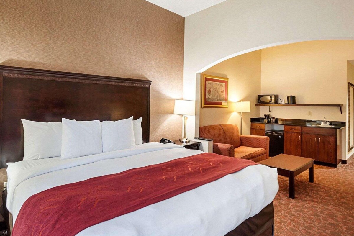 A hotel room with a large bed, a desk, a chair, a kitchenette, and a painting on the wall.