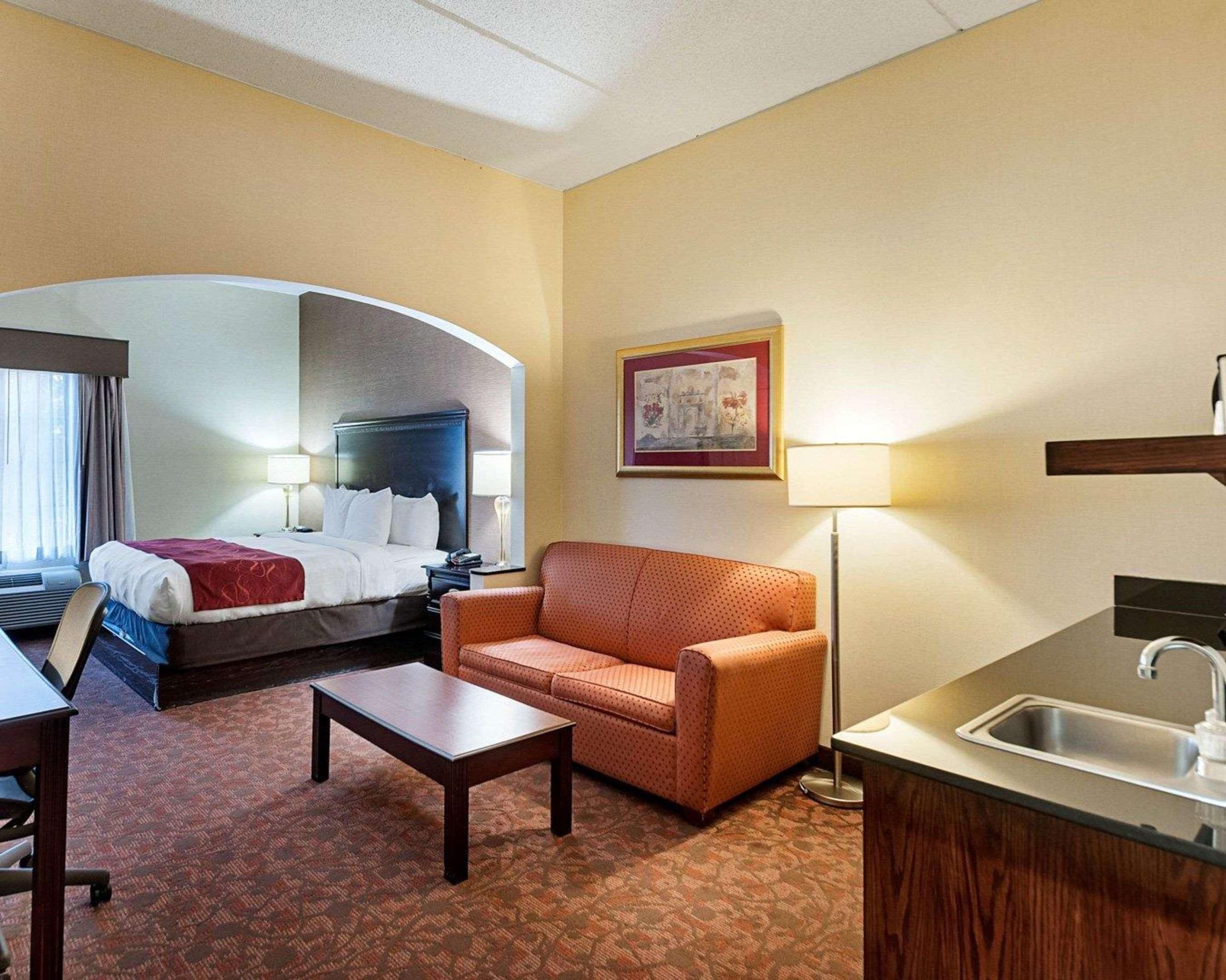 A hotel room with a bed, a sofa, a small table, a desk, a chair, a sink, and a framed picture on the wall.