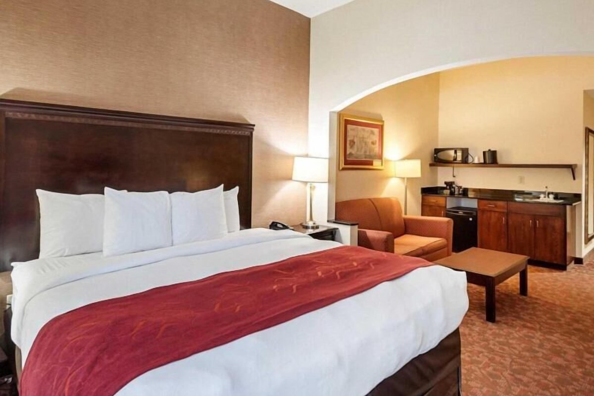 A hotel room with a large bed, a desk, a chair, a kitchenette, and a painting on the wall.