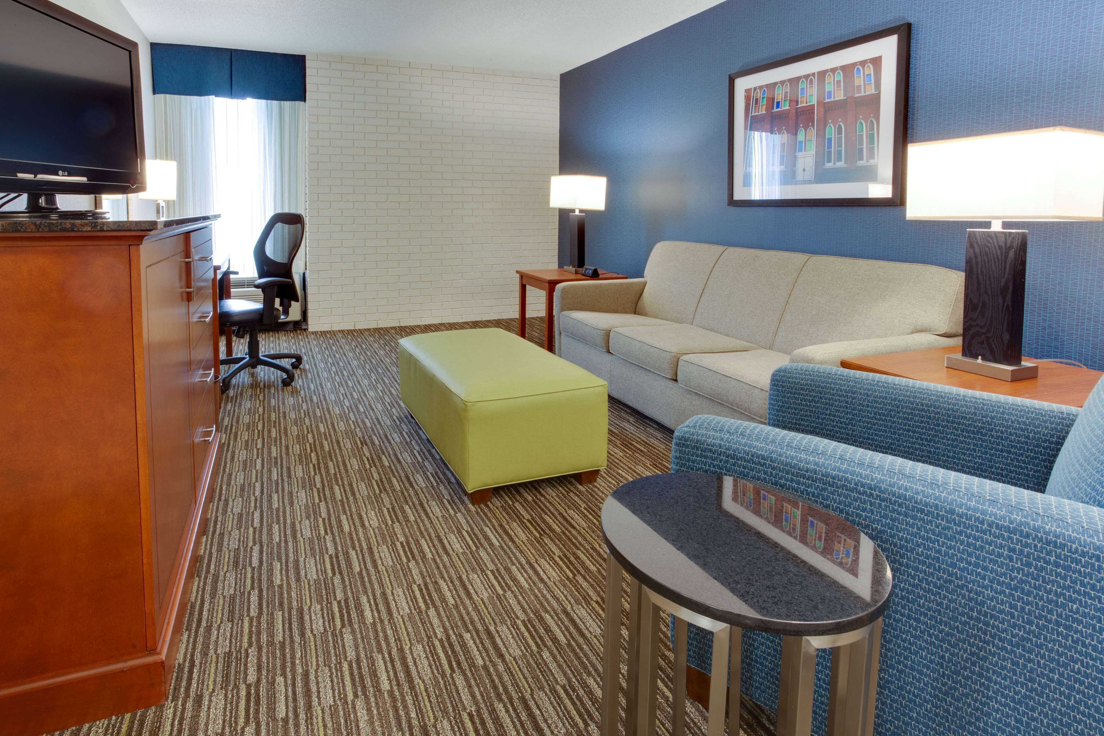 Habitación de hotel con televisión de pantalla plana, un sofá, una pequeña mesa, un reposapiés amarillo, una silla azul estampada y un cuadro en la pared.