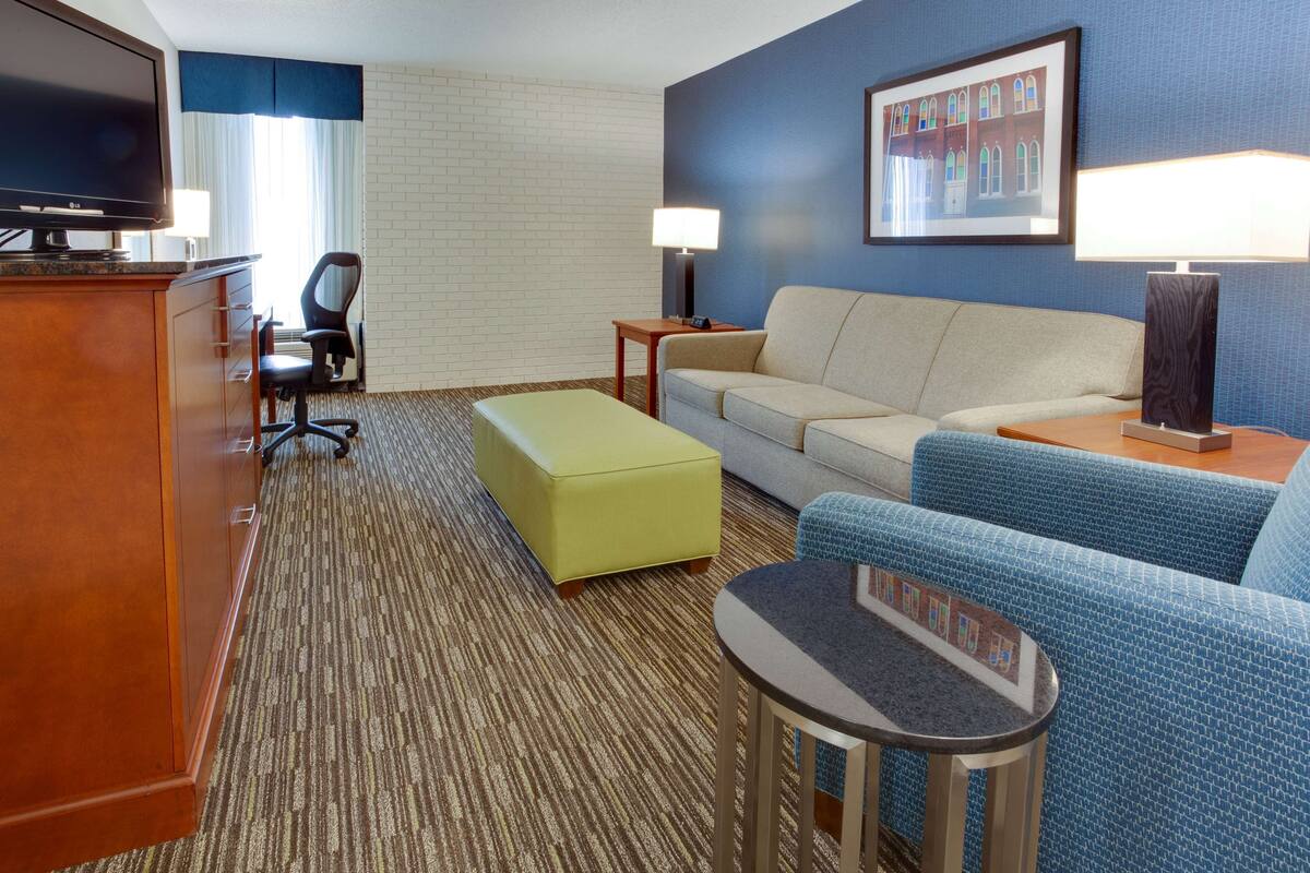 A hotel room with a flat-screen TV, a sofa, a small table, a yellow ottoman, a blue patterned chair, and a framed picture on the wall.