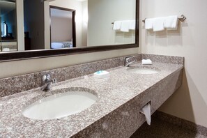Baño de hotel con dos lavamanos, un espejo grande y un perchero con varias toallas.