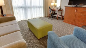 A hotel room with a sofa, an ottoman, a desk with a chair, a television, and a cabinet.