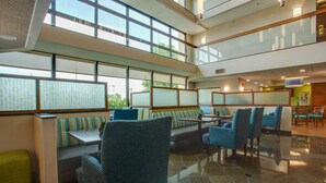 Un moderno vestíbulo de hotel con techos altos, amplias ventanas y diversas opciones de asiento, incluyendo cabinas y mesas.