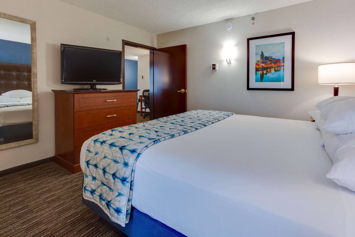 A hotel room with a bed, a television on a wooden dresser, a door, a framed picture on the wall, and a lamp.
