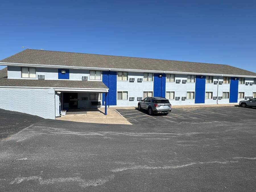 Un motel con una estructura de un piso, zona de estacionamiento techada y cielo azul despejado.