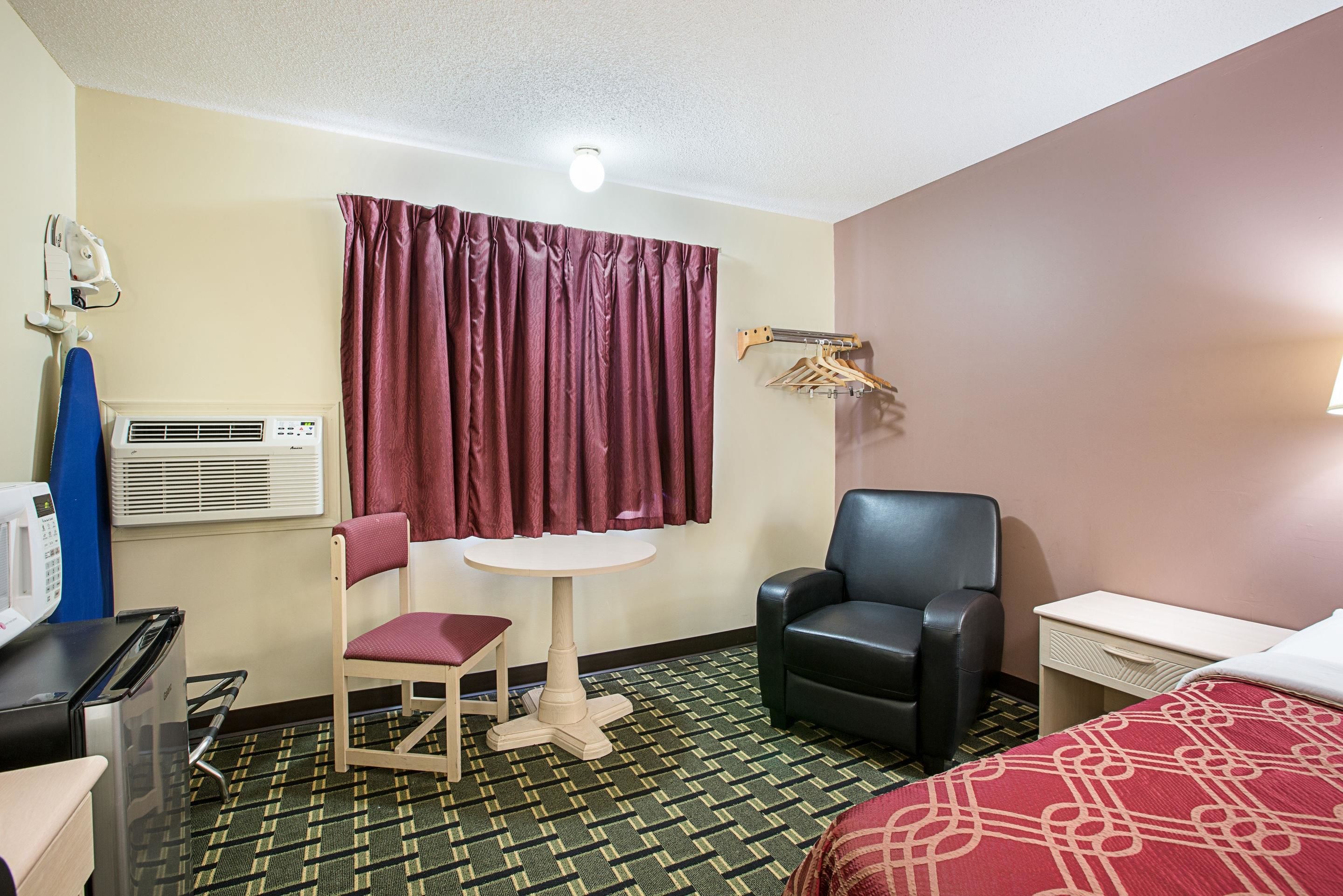 A hotel room with a bed, a desk, a chair, a small table, an air conditioning unit, a microwave, and a curtain.