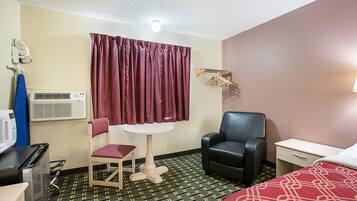 A hotel room with a bed, a desk, a chair, a small table, an air conditioning unit, a microwave, and a curtain.