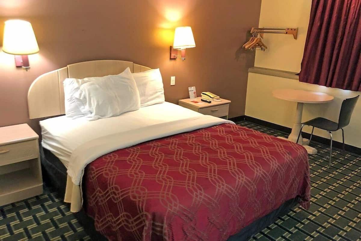 A hotel room with a bed, bedside lamps, a small table, a chair, and a wall-mounted towel rack.