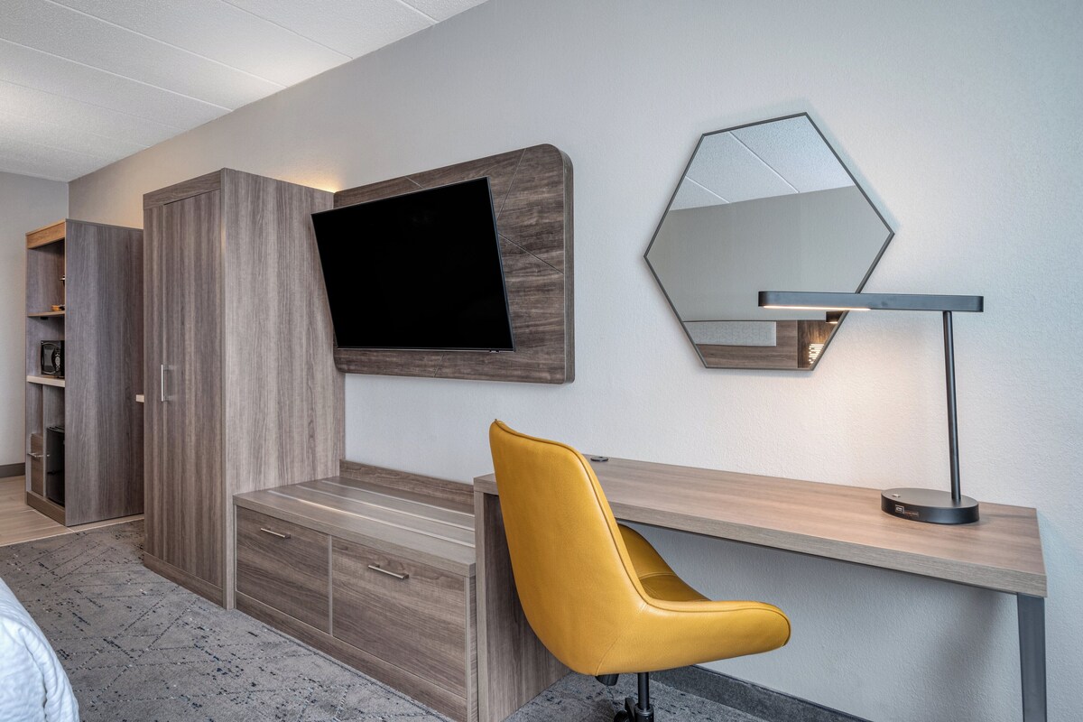Ein modernes Zimmer mit Schreibtisch aus Holz, einem gelben Stuhl, einem Flachbildfernseher und einem Spiegel.
