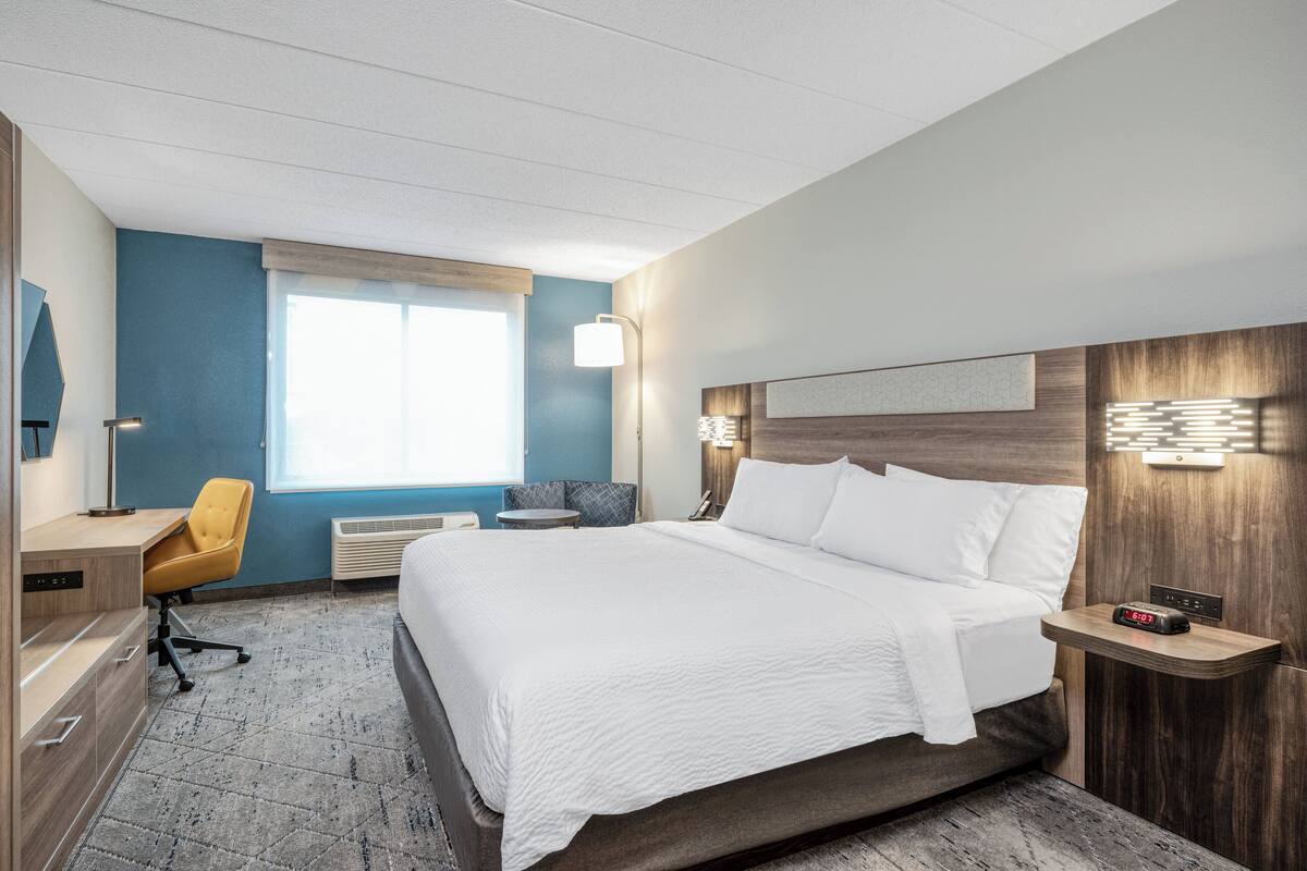 Ein modernes Hotelzimmer mit einem großen Bett, einem Schreibtisch, einem Stuhl und einem Nachttisch mit einer Uhr.