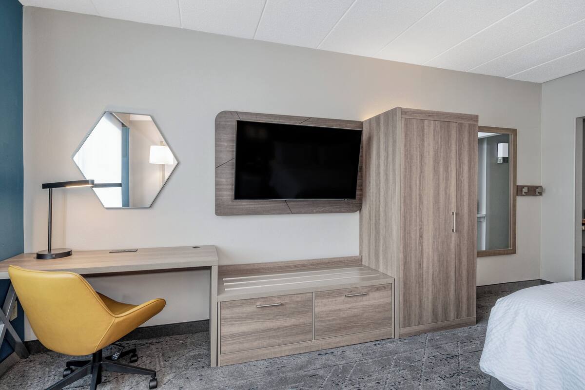 Ein Hotelzimmer mit Schreibtisch, einem gelben Sessel, einem an der Wand befestigten Fernseher, einem Kleiderschrank und einem Bett.