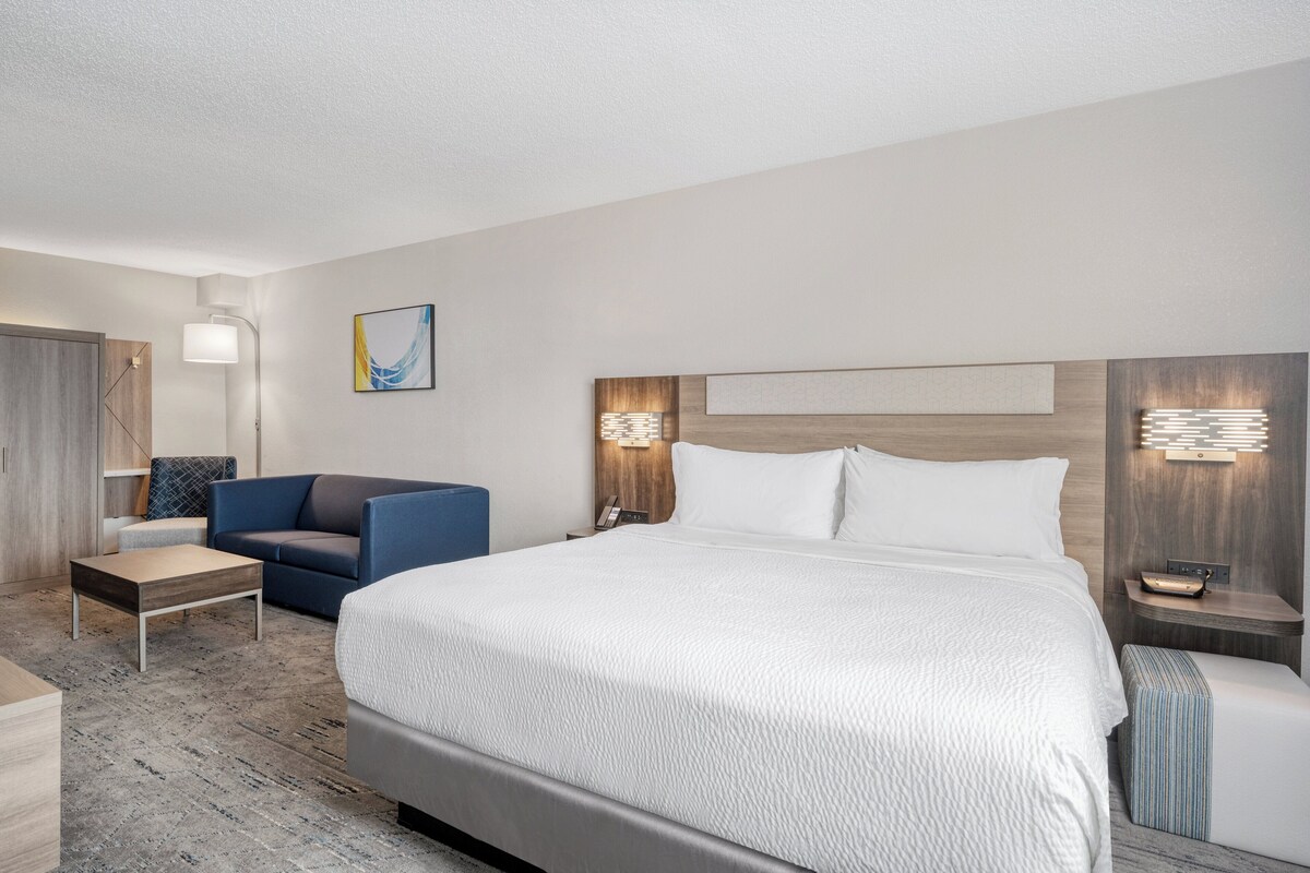 Una habitación de hotel moderna con una cama grande, un sillón azul, una mesita y una mesita de noche con una lámpara.