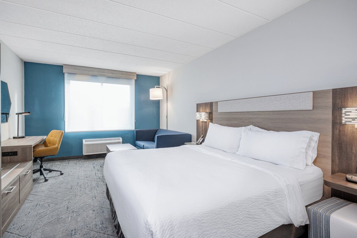 Ein Hotelzimmer mit einem großen Bett, einem Schreibtisch, einem Sessel und einem Fenster mit Jalousien.
