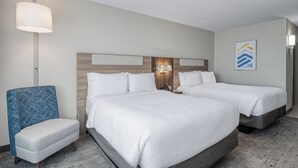 Une chambre d’hôtel avec deux lits, un fauteuil bleu, une table de chevet avec une lampe et une œuvre d’art accrochée au mur.