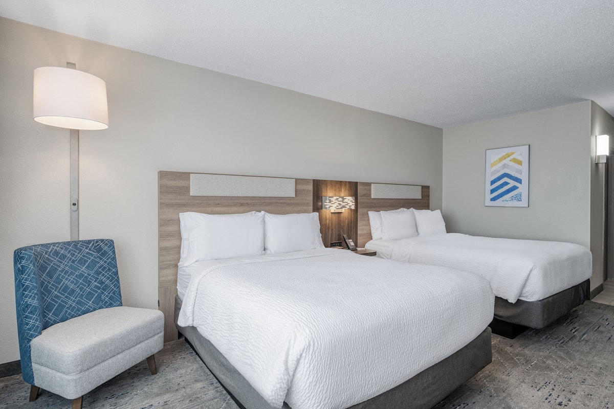 Ein Hotelzimmer mit zwei Betten, einem blauen Sessel, einem Nachttisch mit Lampe und einem Wandbild.