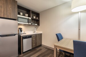 Cocina compacta con microondas, horno y lavavajillas integrados. Hay una mesa de madera con una silla azul y una lámpara.