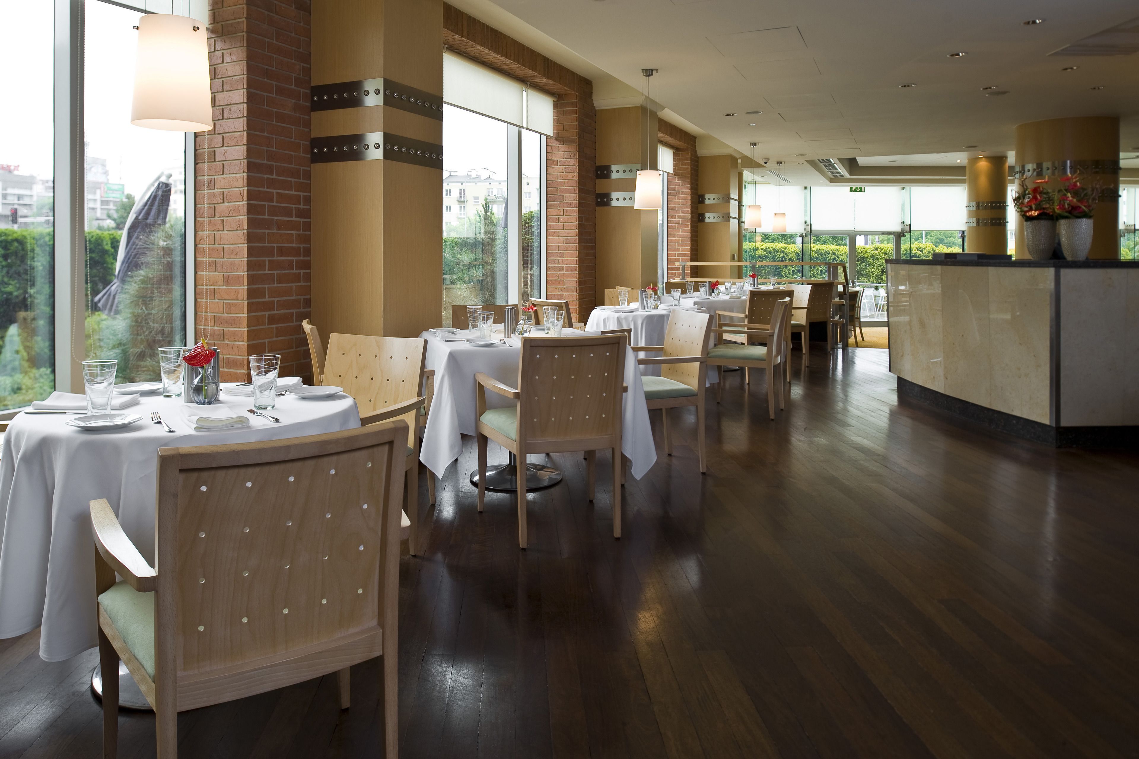 A restaurant with round tables, wooden chairs, and large windows offering a view of greenery.
