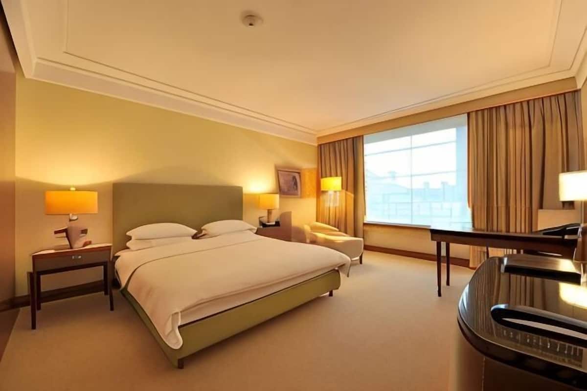 A hotel room with a large bed, two bedside tables with lamps, a desk, a chair, and a view of the city.