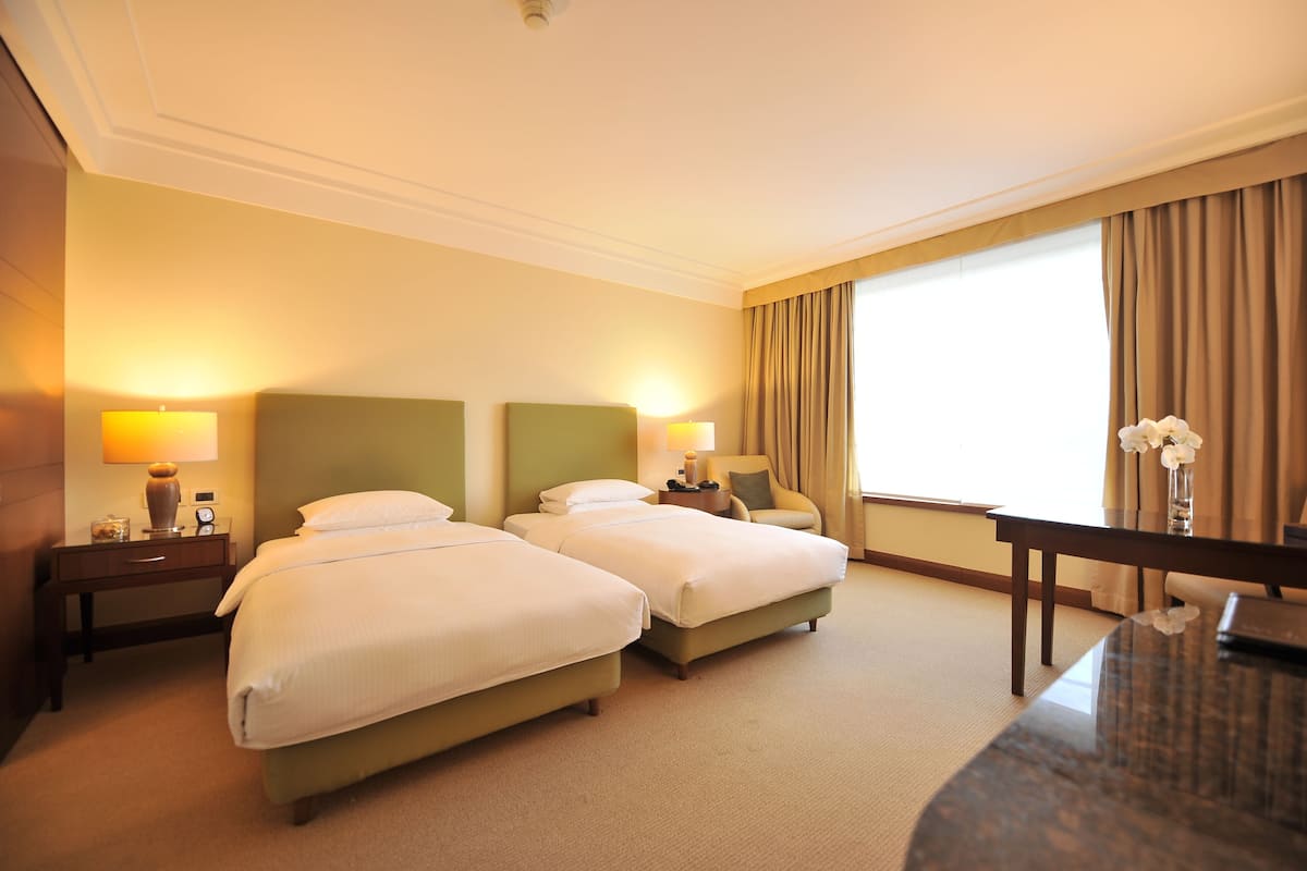 A hotel room with two beds, a desk, a chair, a lamp, and a window with curtains.