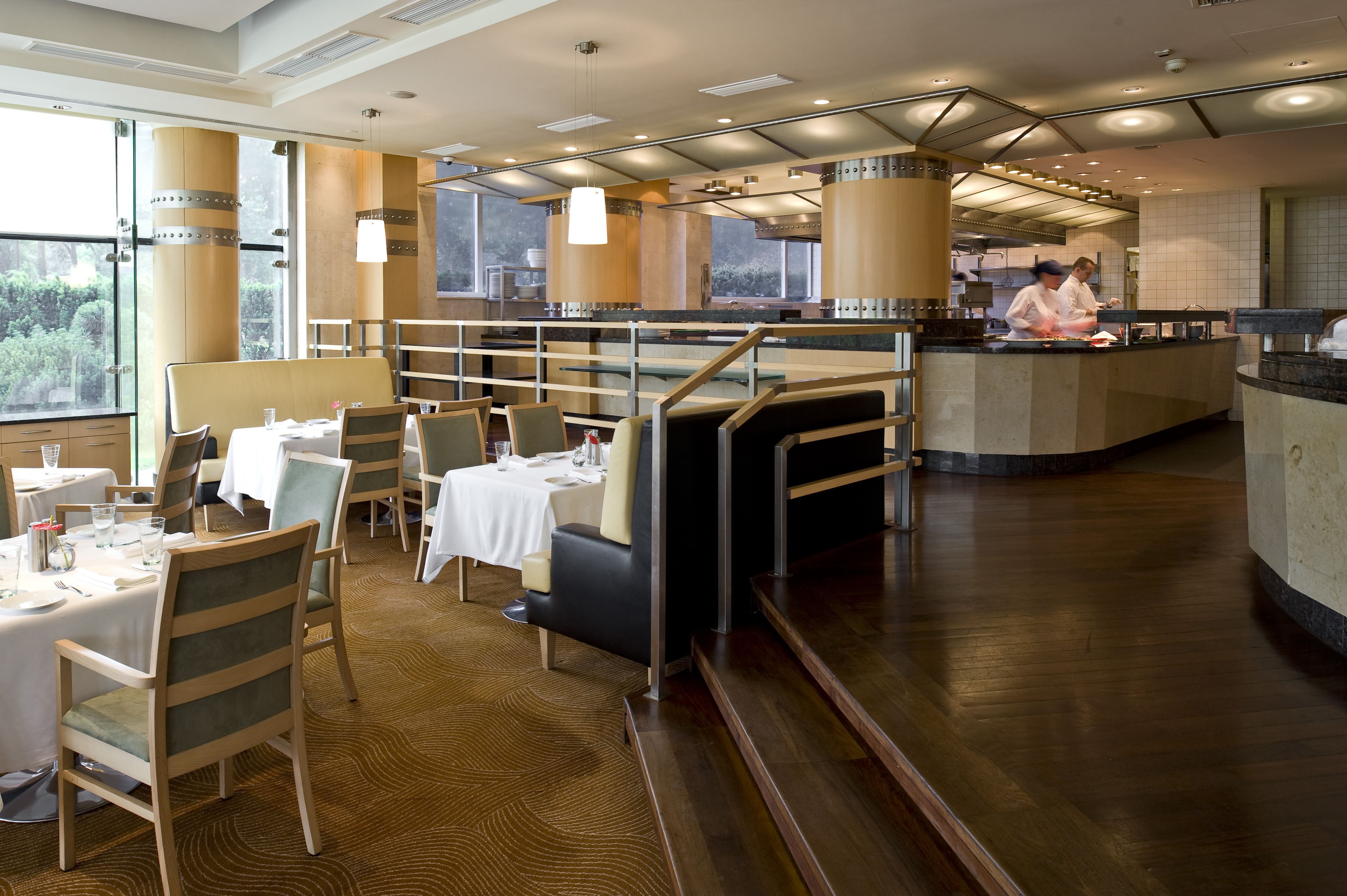 A restaurant with a modern interior, featuring a dining area with tables and chairs, a kitchen area with chefs, and large windows providing natural light.