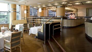 A restaurant with a modern interior, featuring a dining area with tables and chairs, a kitchen area with chefs, and large windows providing natural light.
