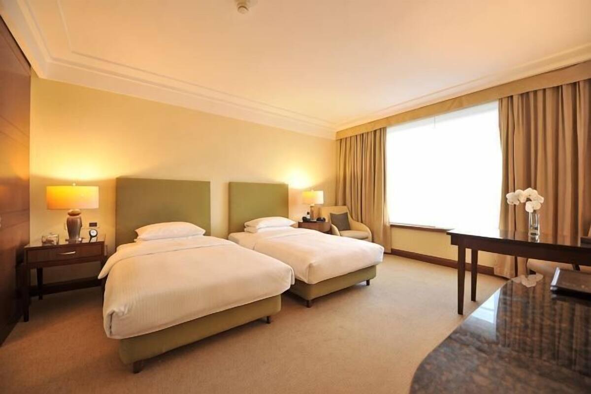 A hotel room with two beds, a desk, a lamp, and a large window.