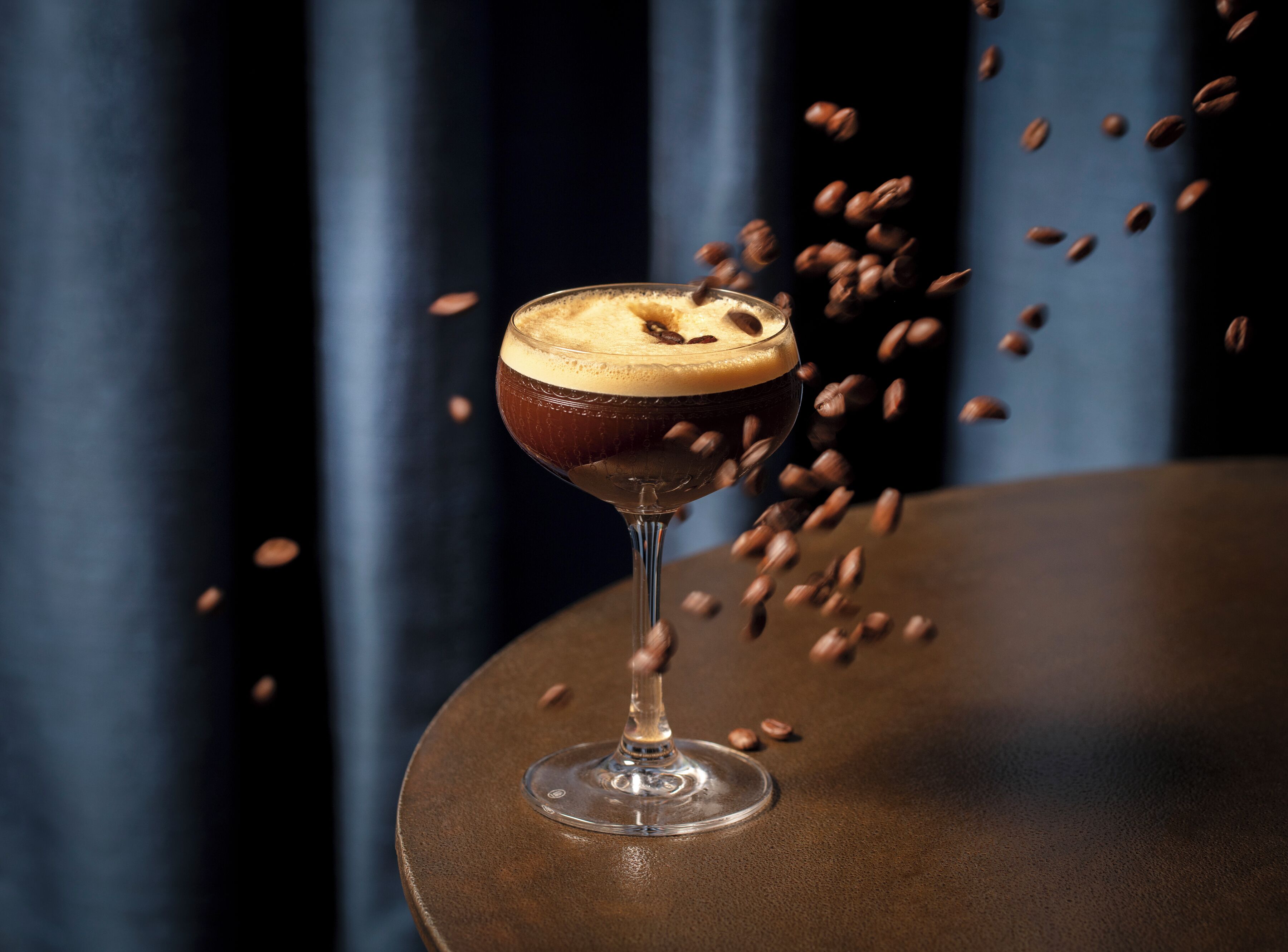 Una copa de cóctel con una espuma en la parte superior, rodeada de granos de café sobre una mesa redonda sobre un fondo oscuro.