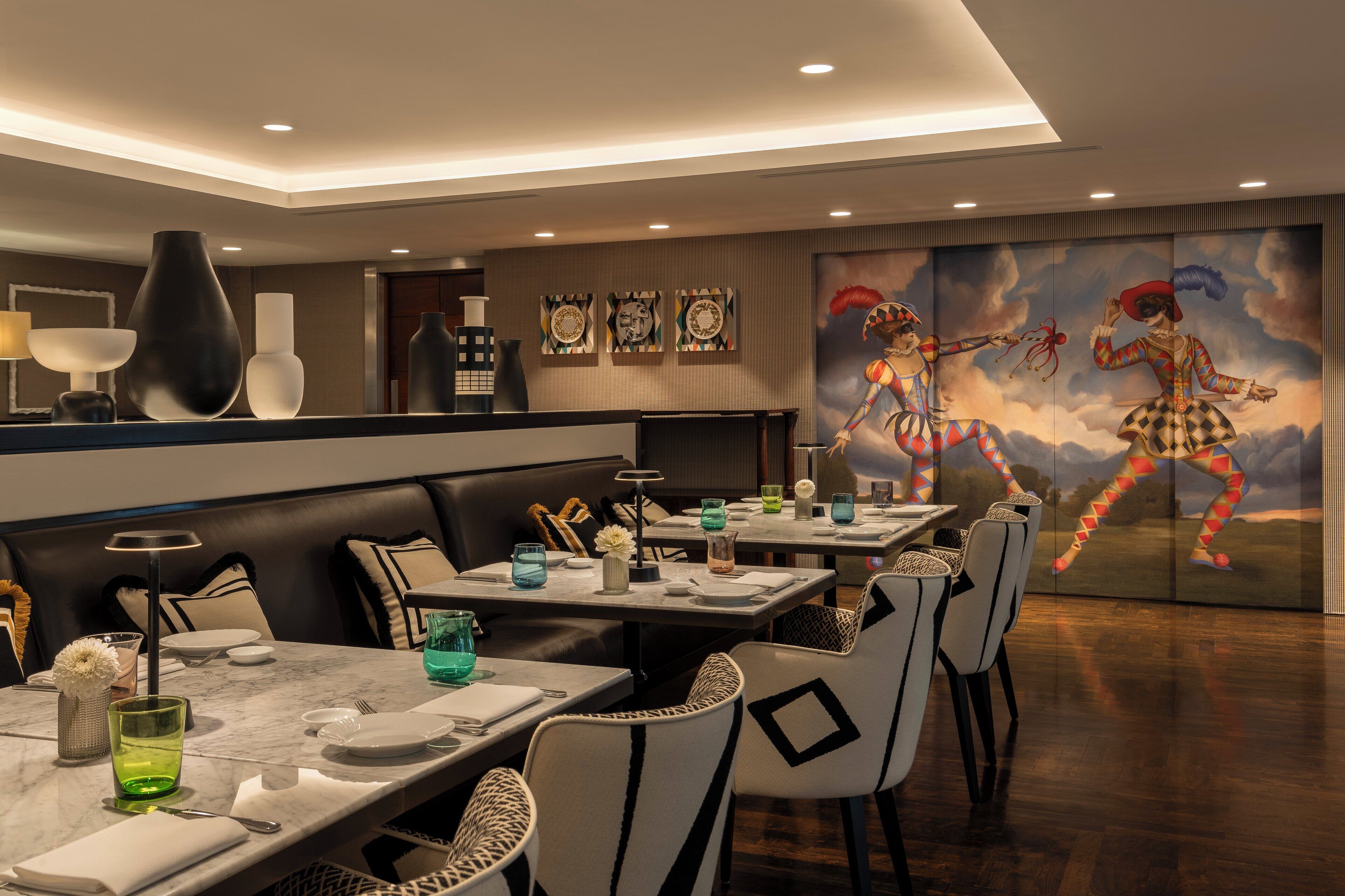 Un restaurante con mesas de mármol, sillas de mimbre y un mural con payasos en la pared.