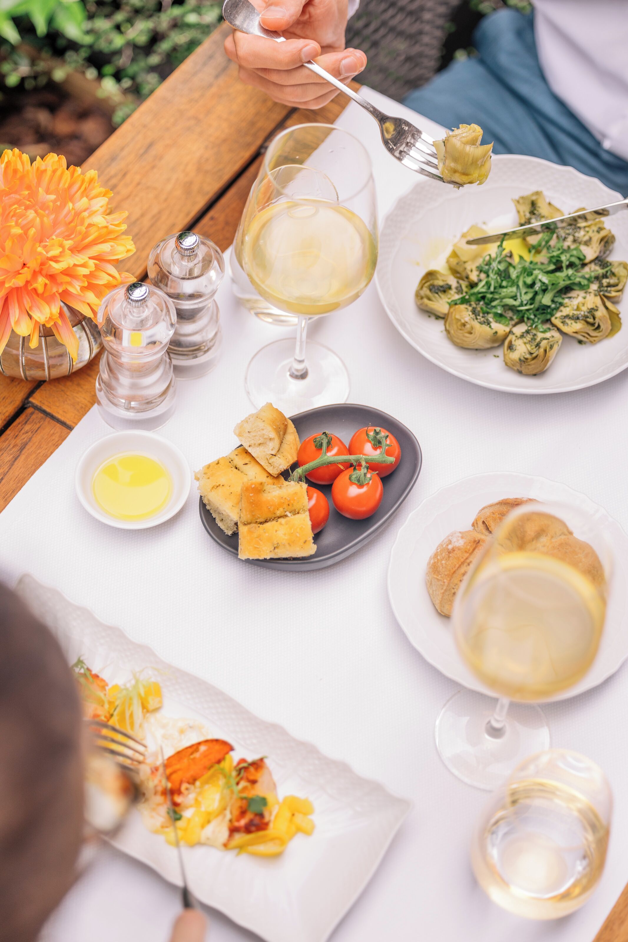 A table set with various dishes, including a plate of artichoke hearts, a plate of cherry tomatoes, and a plate of bread. There are also salt and pepper shakers, a small bowl of butter, and a glass of white wine.