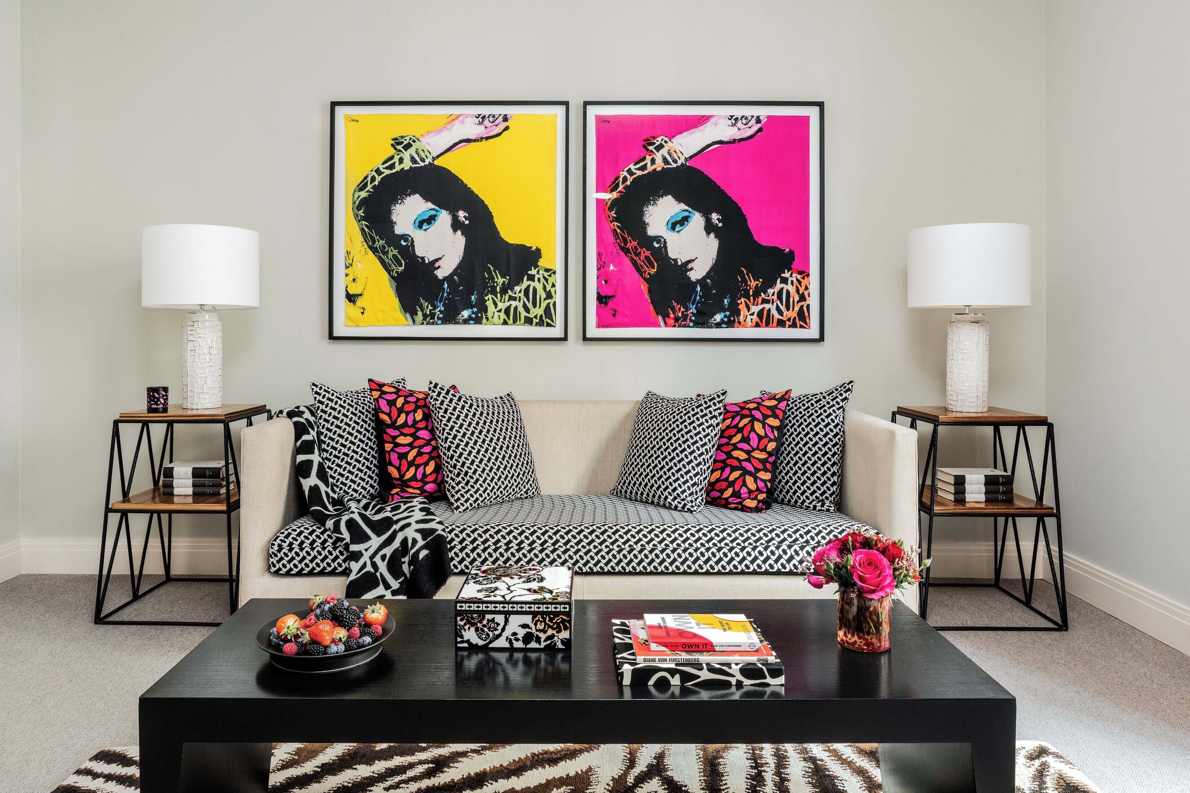 A living room with a sofa, two side tables, a coffee table with books and a fruit bowl, and two framed artworks on the wall.