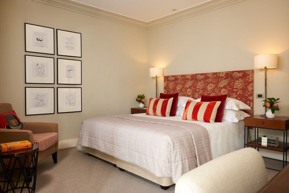 Una habitación de hotel con una cama grande, dos mesitas de noche con lámparas, una silla, una mesita pequeña con un jarrón y cuadros en la pared.