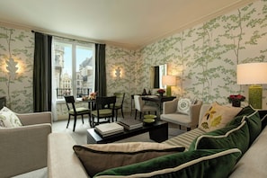 A hotel room with a dining area, a sofa, a coffee table, and a view of the city through the window. - Hotel Amigo, a Rocco Forte Hotel (Brussels)