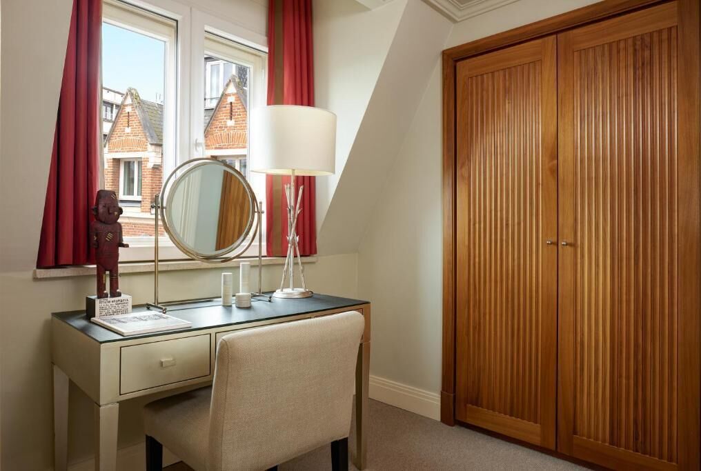 A bedroom with a wooden wardrobe, a desk with a mirror, and a window with red curtains.