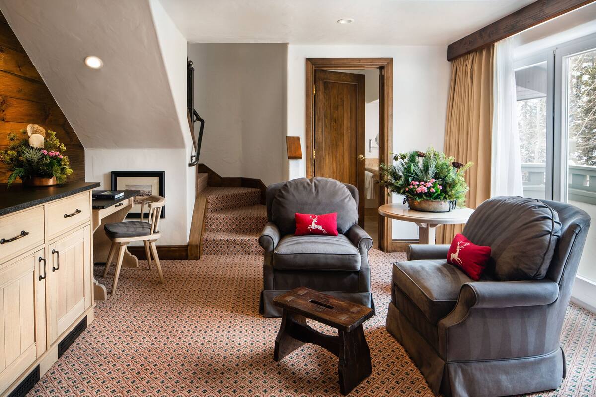 A cozy living room with a fireplace, two leather armchairs with red cushions, a wooden coffee table, and a patterned carpet.