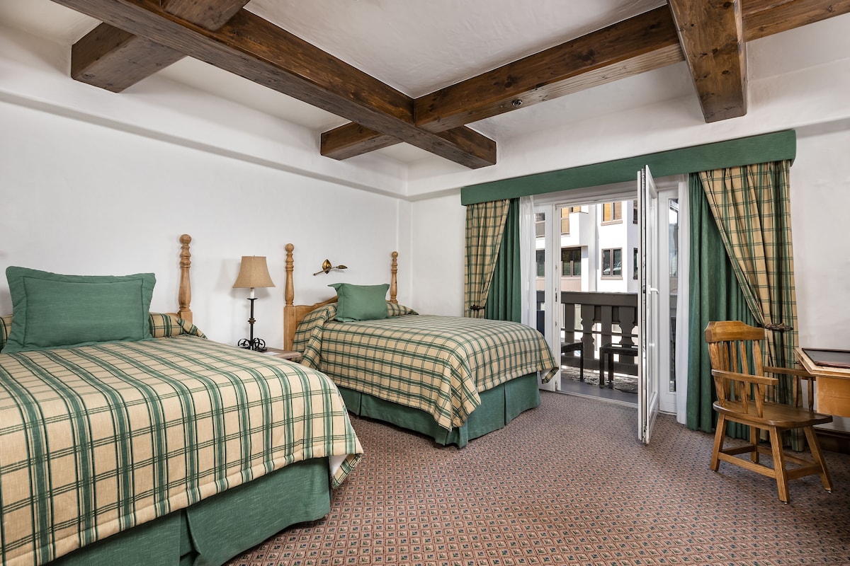 A room with two beds, a wooden chair, a table, and a balcony with curtains.