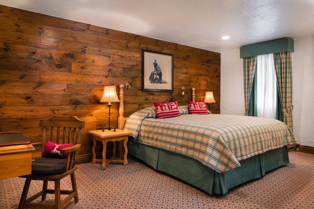 A bedroom with a wooden wall, a bed with plaid bedding, two bedside lamps, a chair, a desk, and a framed picture on the wall.